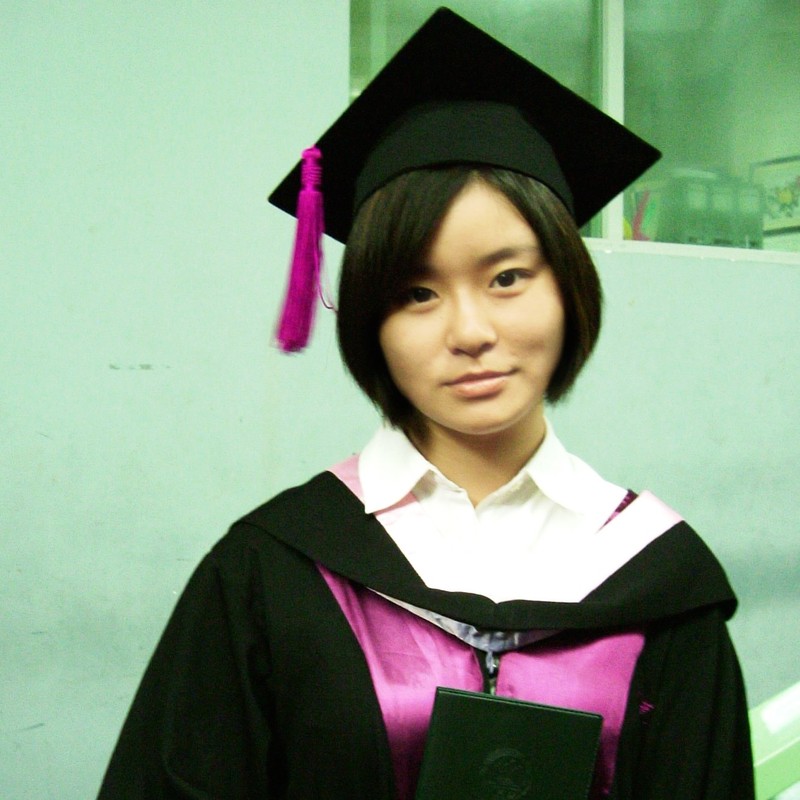 An East Asian woman dressed in a graduation cap and gown wears a neutral or subdued expression. 