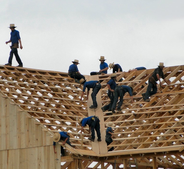 A group of Amish men dressed in traditional clothes and hats work together to build a barn.