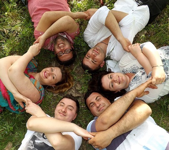 A group of friends lay together on the grass in a circle holding hands.
