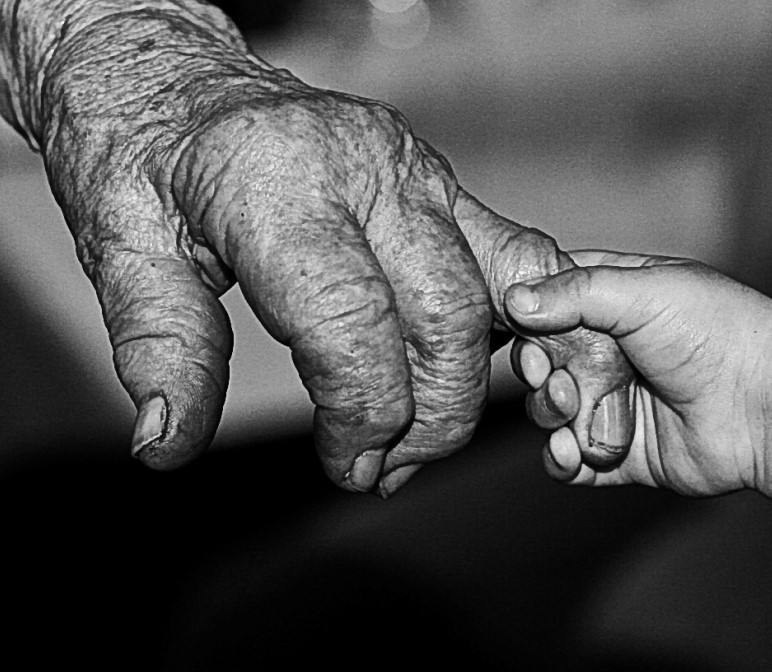 A very small hand of a child holds a wrinkled finger on the hand of a very old person.