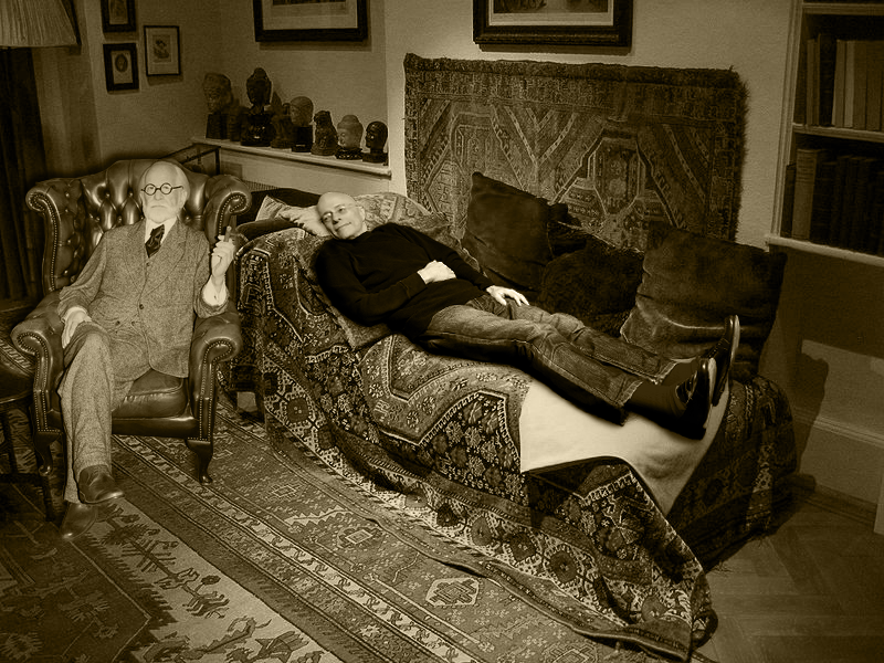 This is a black and white image of Sigmund Freud conducting psychoanalysis with a patient. The image shows Freud, wearing a suit and glasses, sitting in a leather chair facing away from the patient. The patient, a middle-aged man, is lying on a couch that is covered by a patterned blanket that is similar to a number of oriental rugs covering the floor. A number of framed photographs and books can be seen in the background.